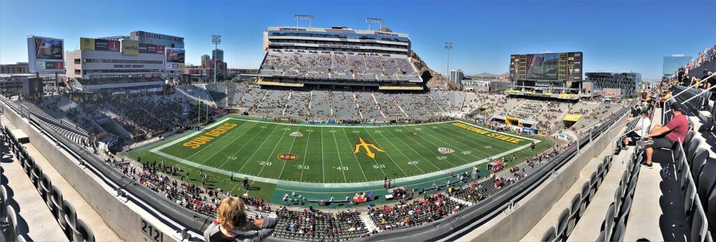 A panoramic view across a packed college football stadium in Tempe with the field, stands and scoreboards under a blue sky
