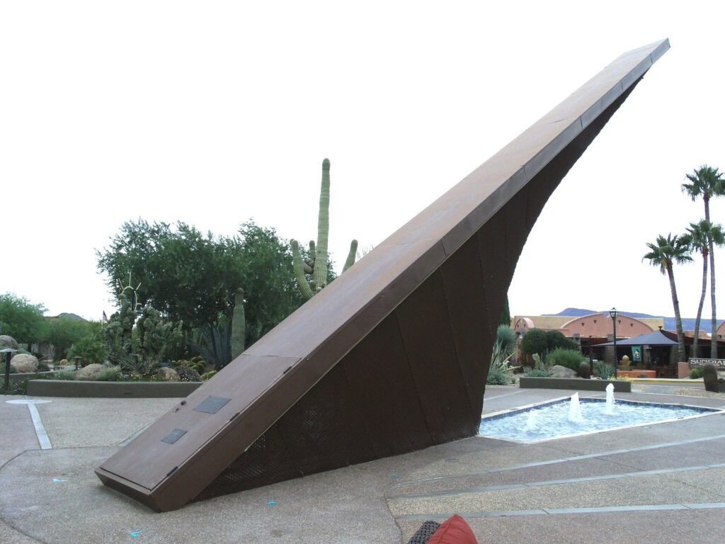 The Carefree Sundial's long, angled arm rising over a small fountain pool, with saguaros and palms behind