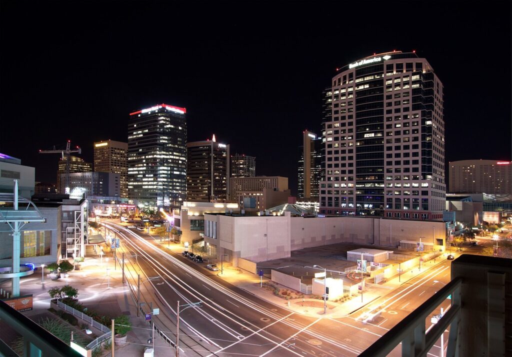 Downtown Phoenix high-rises lit up at night above a wide street with light-rail tracks and streaks of car headlights