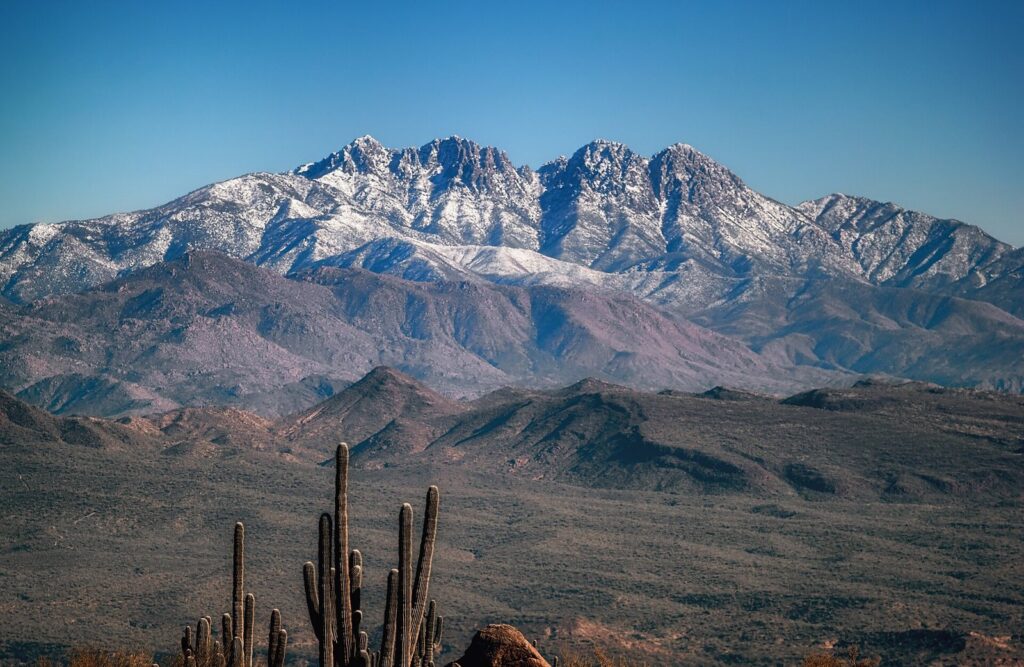 Snow on the jagged summits of Four Peaks above desert ridges, with saguaro cacti in the foreground under a blue sky