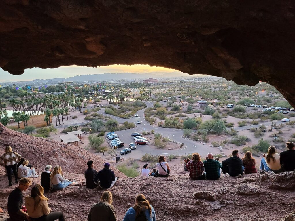 People sitting inside the sandstone opening of Hole-in-the-Rock at Papago Park, looking out over palms, a parking lot and the Phoenix skyline at dusk