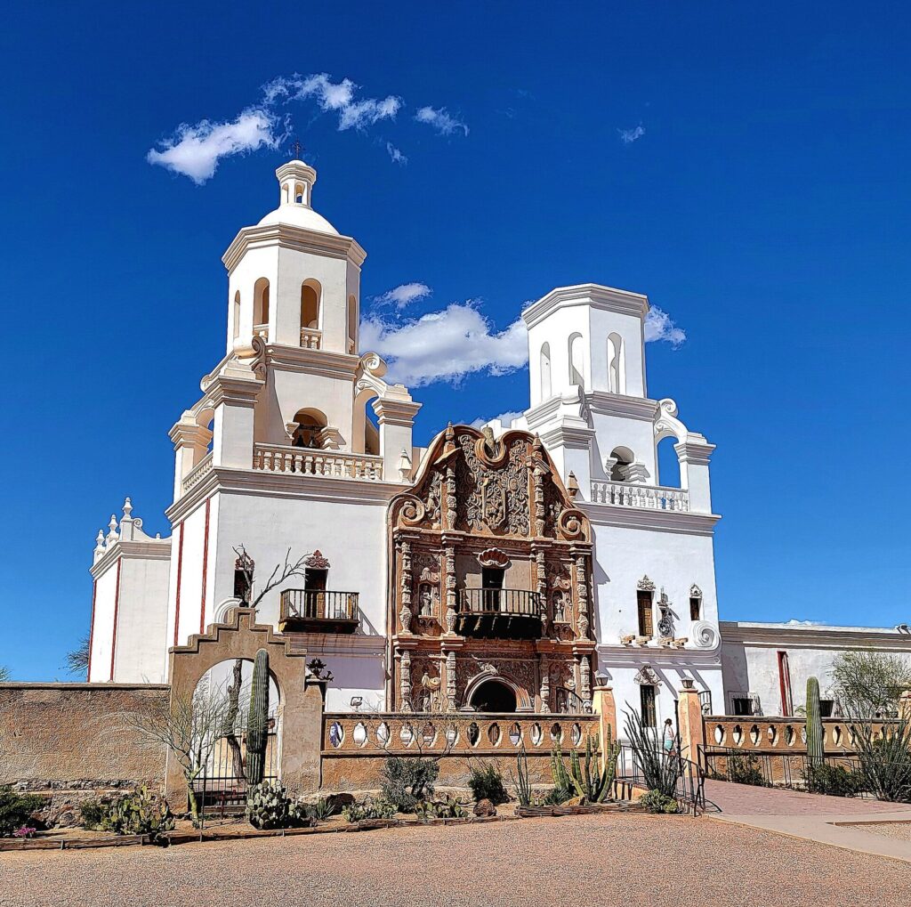 The white facade and twin bell towers of Mission San Xavier del Bac under a deep blue sky, with a carved brown entrance at the center