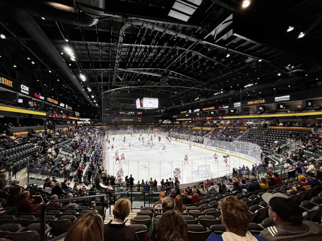 Fans in the stands watching a hockey game on the brightly lit ice inside Mullett Arena in Tempe