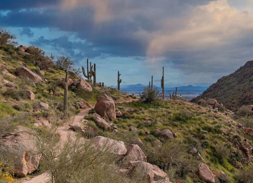 A rocky trail winding past saguaros and granite boulders on Pinnacle Peak, with storm clouds over the Valley beyond
