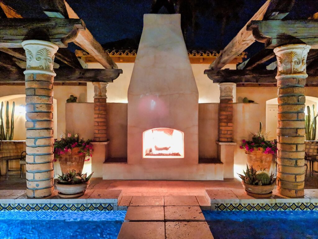 A lit outdoor fireplace between stone columns at the edge of a tiled pool at Royal Palms Resort at night