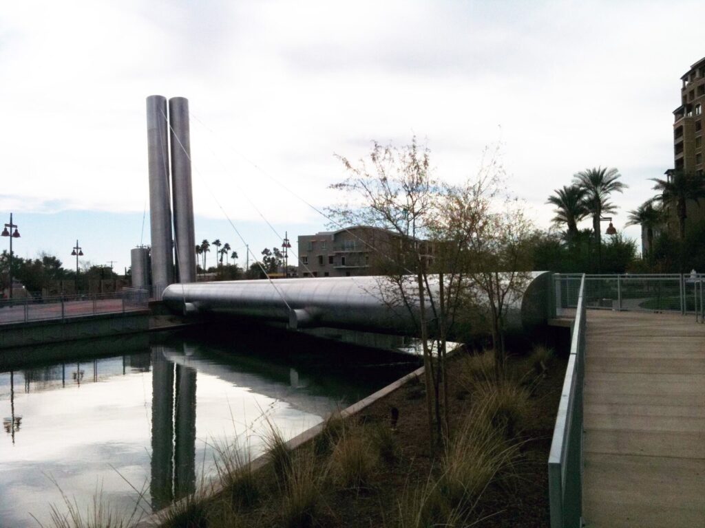 The Soleri Bridge's two tall steel pylons beside the Arizona Canal at the Scottsdale Waterfront, with trees and a wooden walkway
