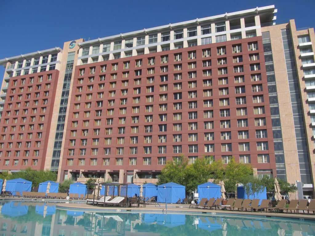A resort pool lined with blue cabanas below a tall red-and-tan hotel tower