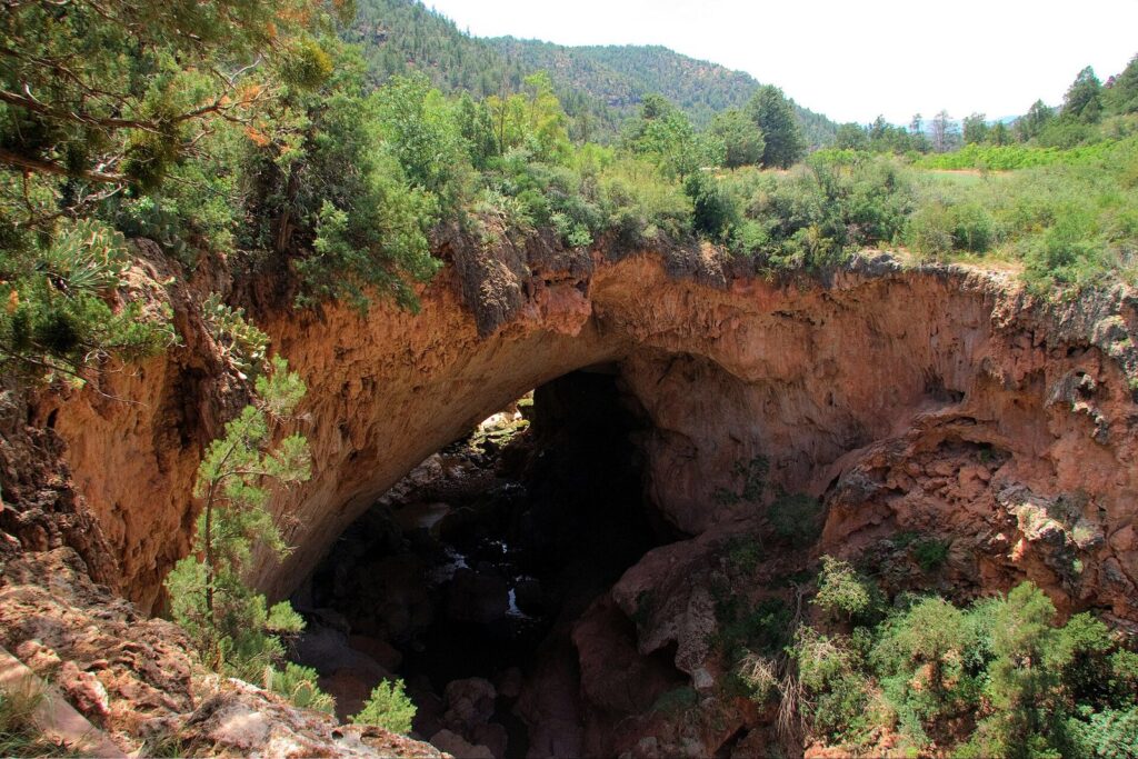 A massive rust-colored travertine arch over a shaded creek, surrounded by green trees and brush at Tonto Natural Bridge State Park