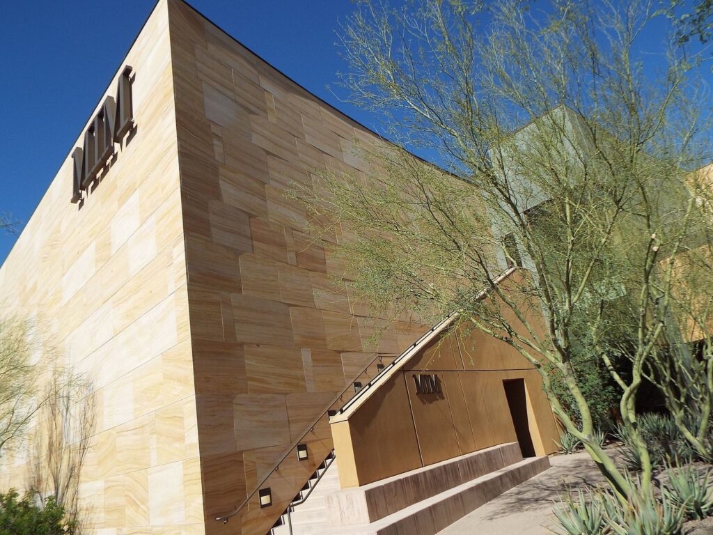 The curved sandstone-colored wall of the Musical Instrument Museum under a blue sky, with desert trees in front