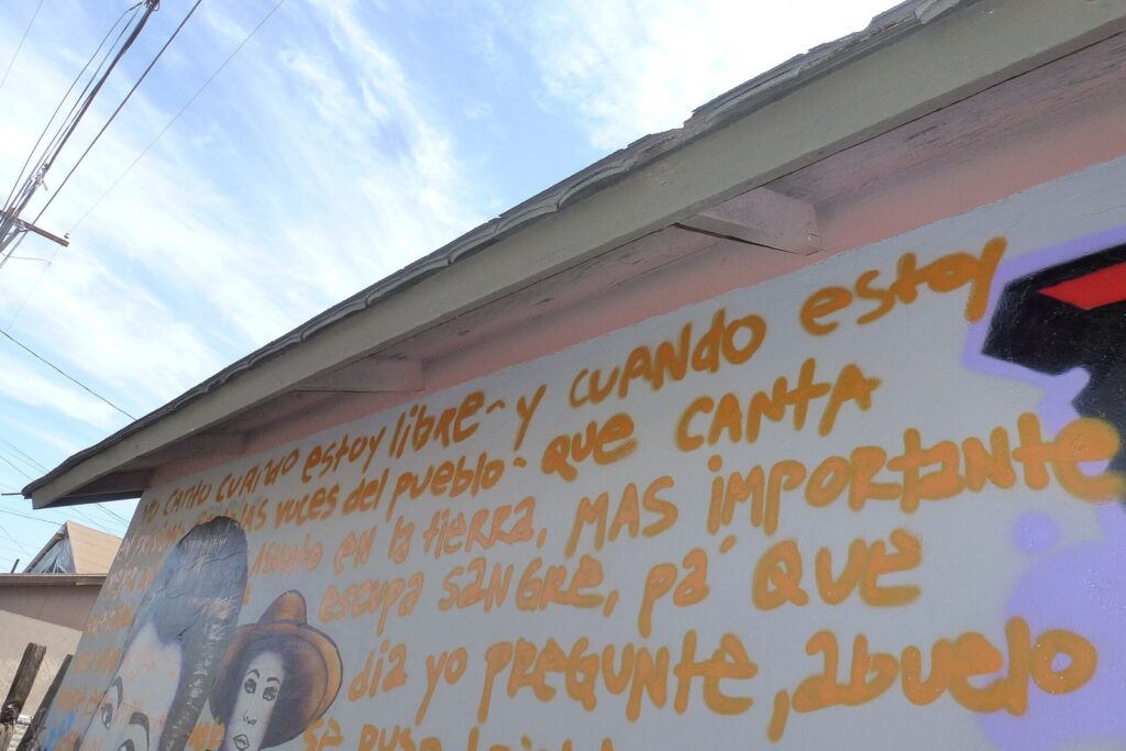 A mural of painted faces and Spanish-language lettering on the side of a low building on Roosevelt Row