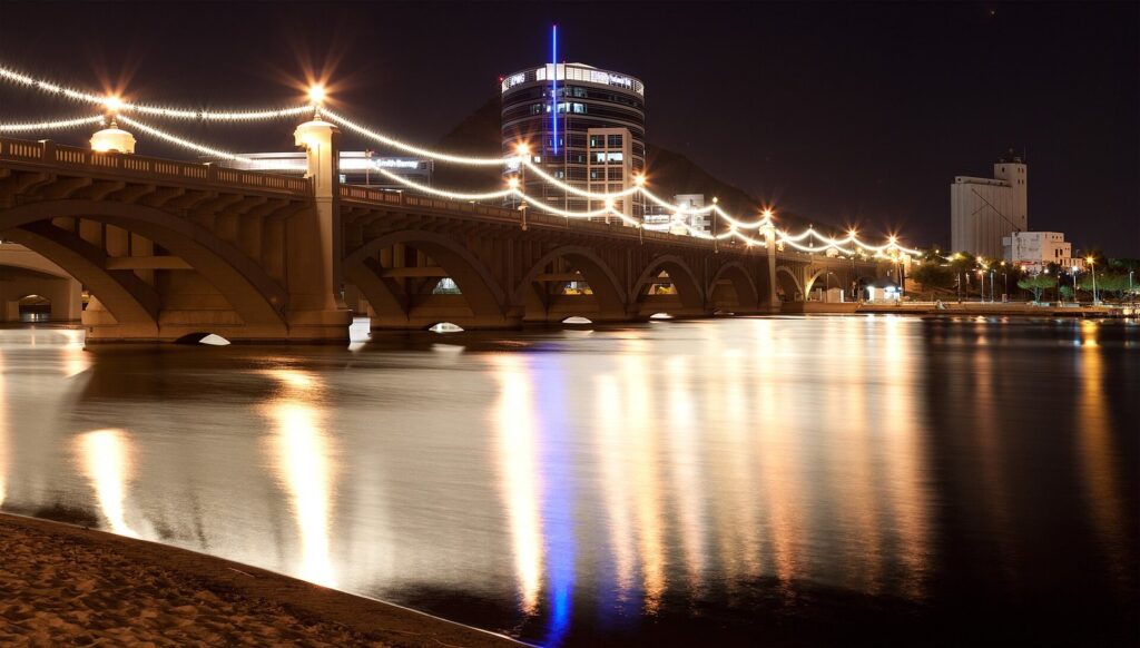 The lit arches of the Mill Avenue bridge reflected in Tempe Town Lake at night, with office buildings beyond